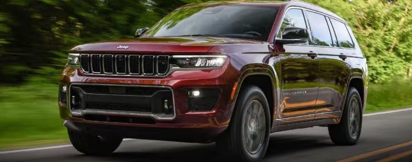 A maroon 2023 Jeep Grand Cherokee is shown driving on a tree-lined road.
